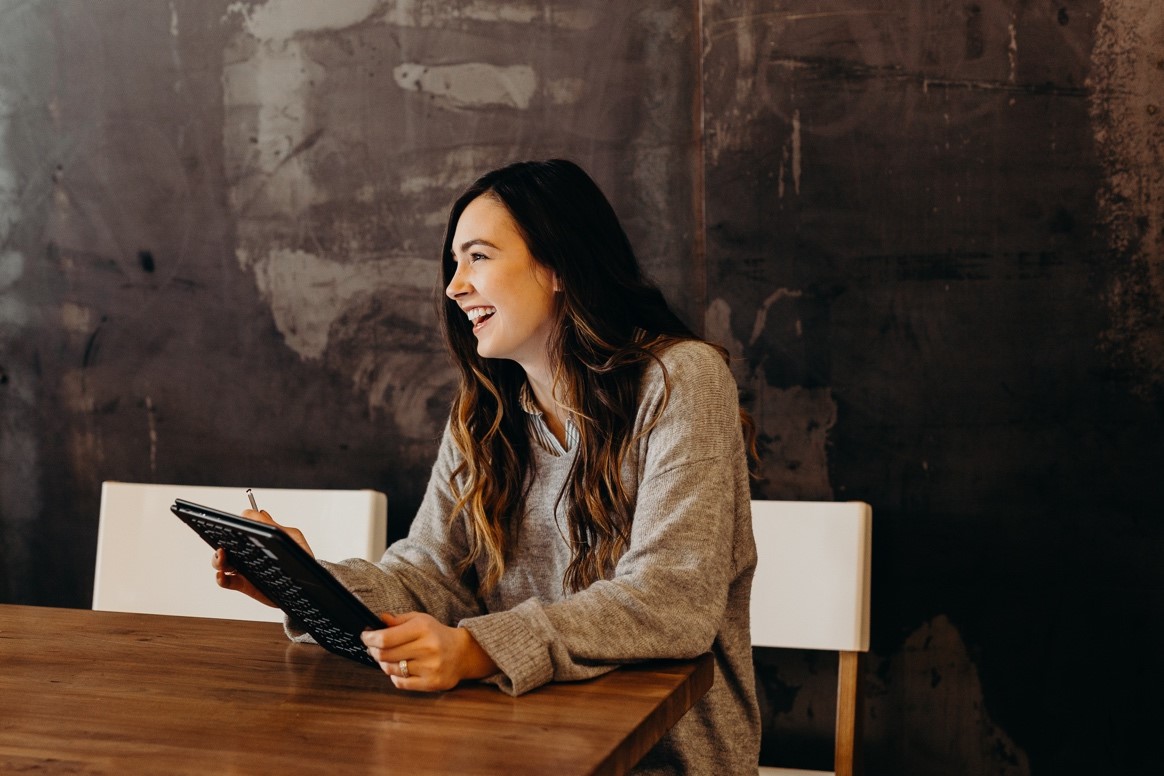 A woman seated at a table, smiling while using a tablet, exuding a sense of joy and engagement in her activity.